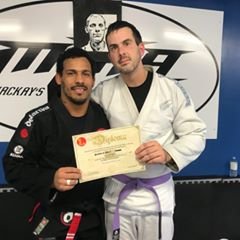 Two men in martial arts uniforms holding a diploma, standing in front of a wall with a martial arts logo.