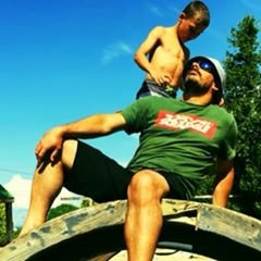 Man wearing sunglasses and green t-shirt sitting outdoors with a young boy standing behind him on a wooden structure against a blue sky.