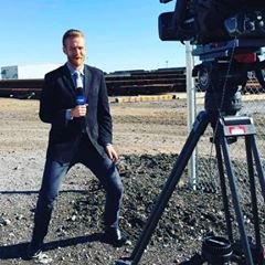 Male news reporter in a suit holding a microphone, standing outdoors on a gravel surface with a large video camera in the foreground.