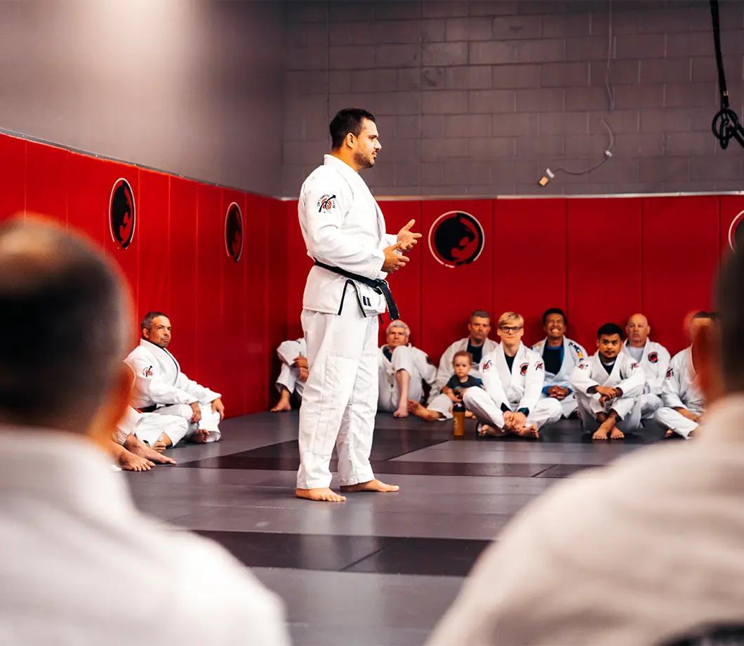 Martial arts instructor in white gi demonstrating or speaking in front of seated students on mats inside a dojo with red padded walls.