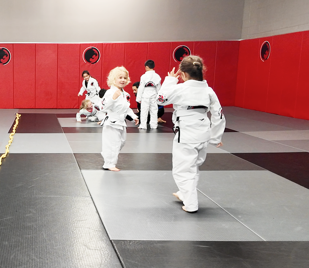Young children in white martial arts uniforms practicing on mats in a dojo with red padded walls.