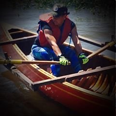 Person wearing a life jacket and gloves rowing a wooden boat on water.