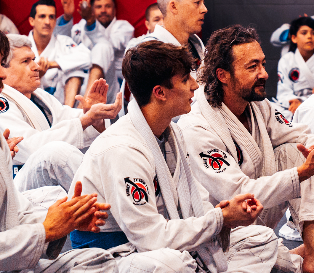 Group of people sitting and clapping while wearing white martial arts uniforms with patches.