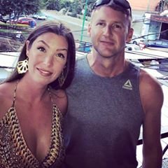 Smiling woman in leopard print dress and man in gray sleeveless shirt posing outdoors near water.