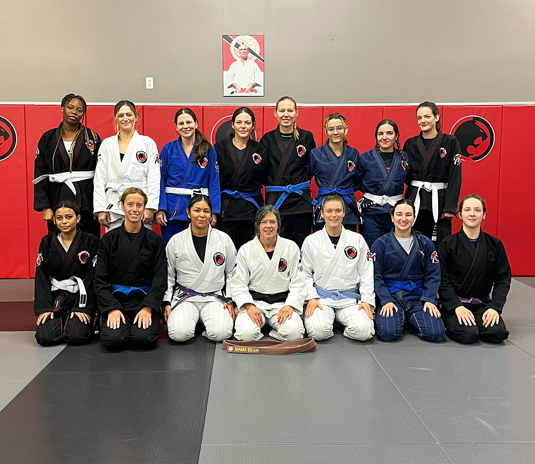 Group of sixteen women martial artists in gis with various belt colors posing in a gym with red padded walls and a poster above.